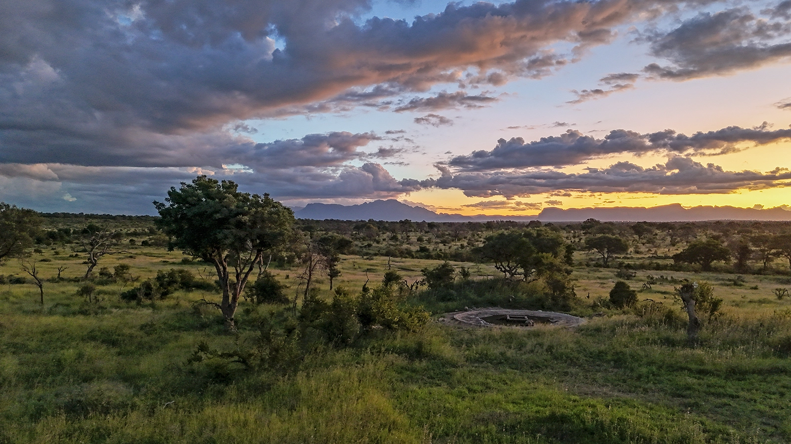 A dramatic African sunset over the open bushveld, with storm clouds lit in shades of orange and purple above a wide savanna landscape and distant mountain silhouettes.