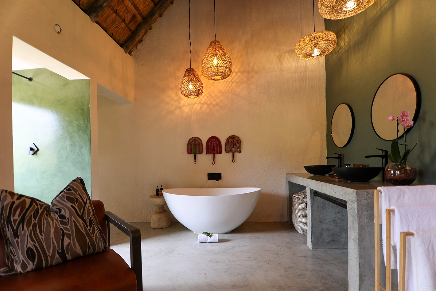 A spacious ensuite bathroom featuring a freestanding white oval bath, concrete double vanity with black basin sinks, round mirrors, woven pendant lights, and African decorative wall fans beneath a thatched ceiling.