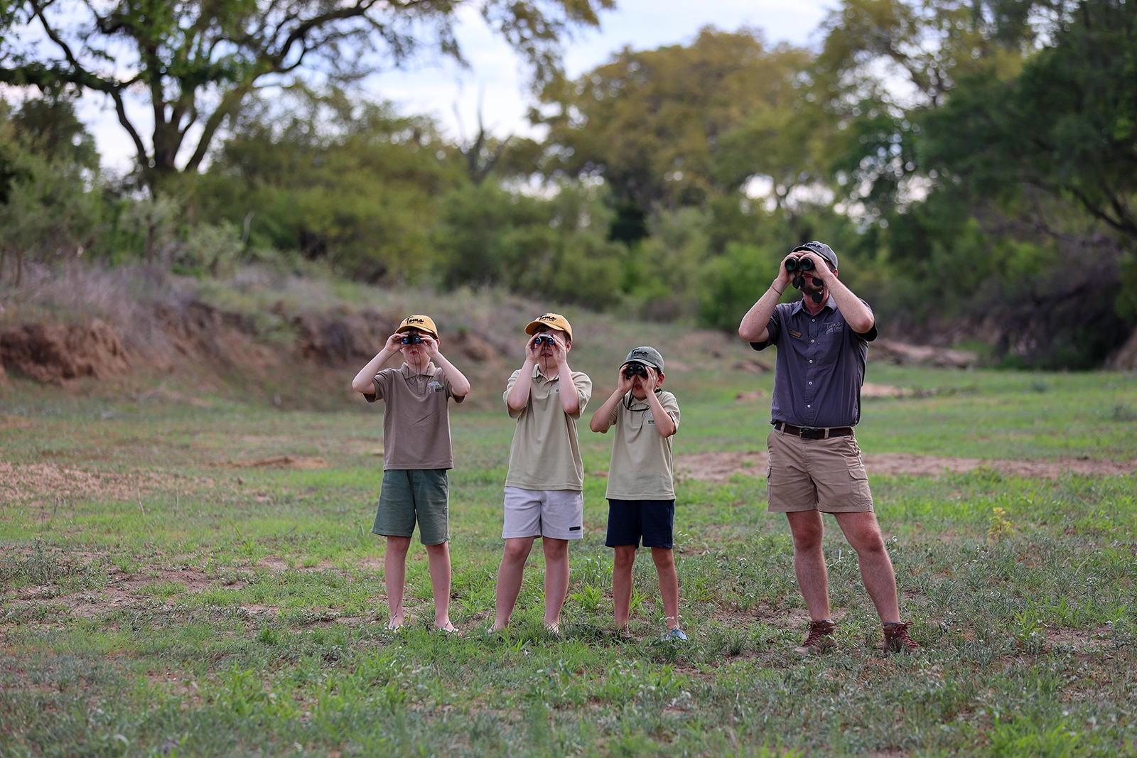A guide and three children stand in an open grassy clearing, all looking through binoculars during a guided safari activity at Tomo Safari Lodge.