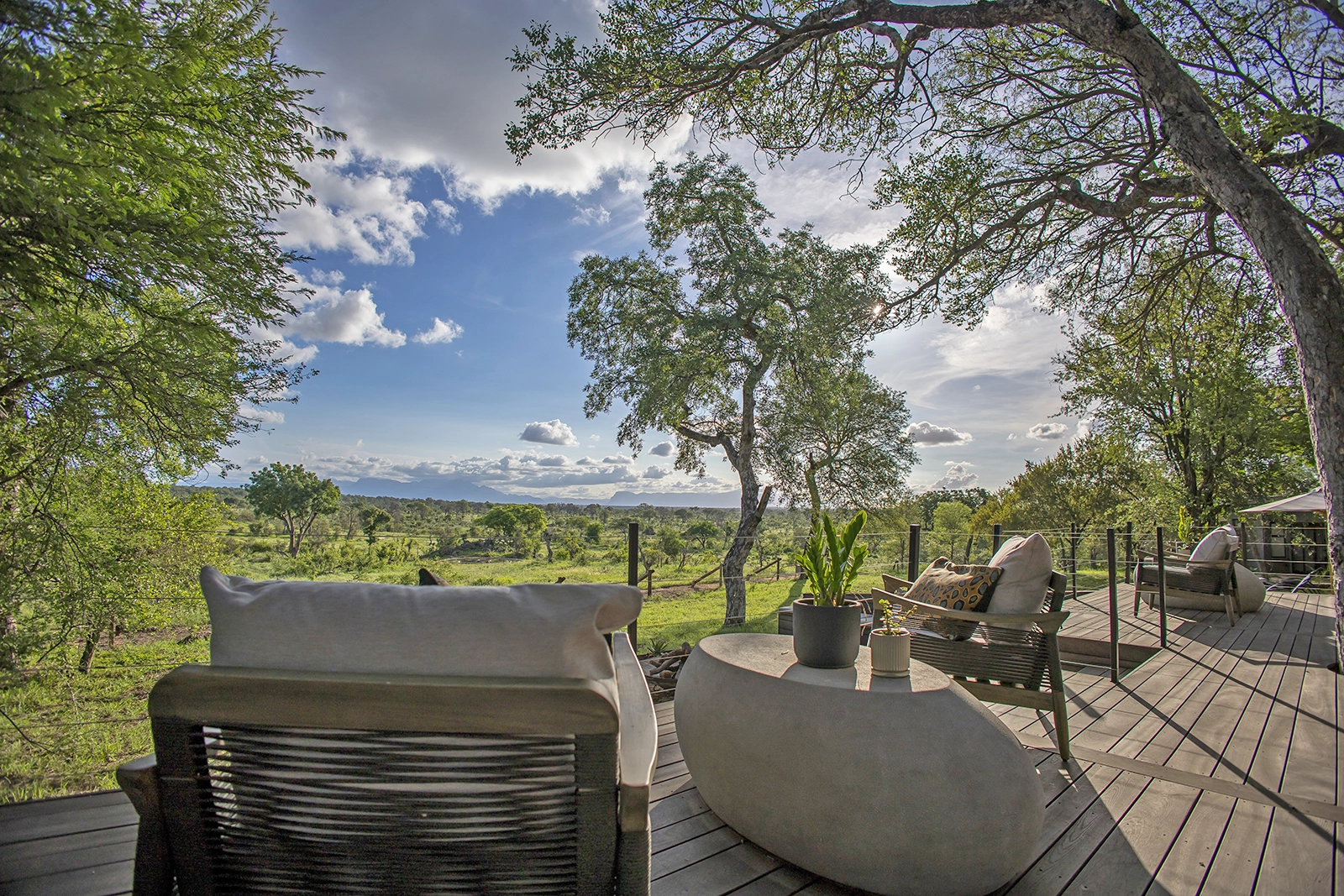 A stylish outdoor deck at Tomo Safari Lodge, furnished with modern lounge chairs and a concrete coffee table, overlooking a lush green bushveld landscape under a blue sky with scattered clouds.