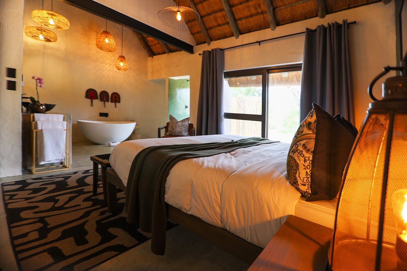 A warmly lit family room interior at Tomo Safari Lodge with a king bed dressed in white linen and a green throw, decorative pillows, a patterned rug, lantern bedside light, thatched roof, and a freestanding bath visible in the open ensuite beyond.