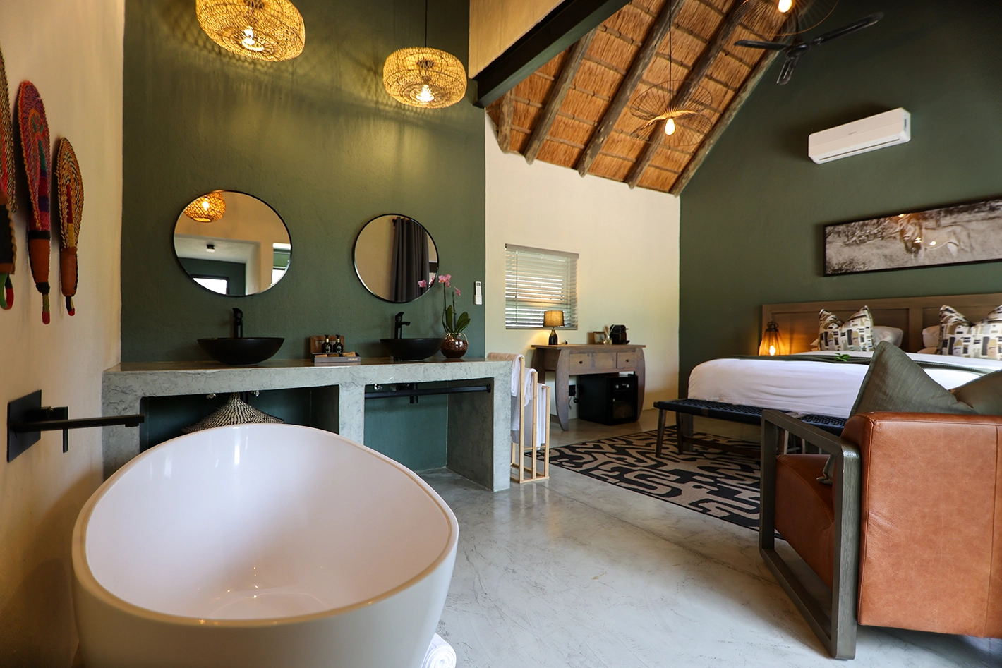 An open-plan view of the Tomo Safari Lodge family room showing the freestanding bath and double vanity in the foreground, leading through to a king bed, patterned rug, leather armchair, and thatched ceiling with woven light fittings.
